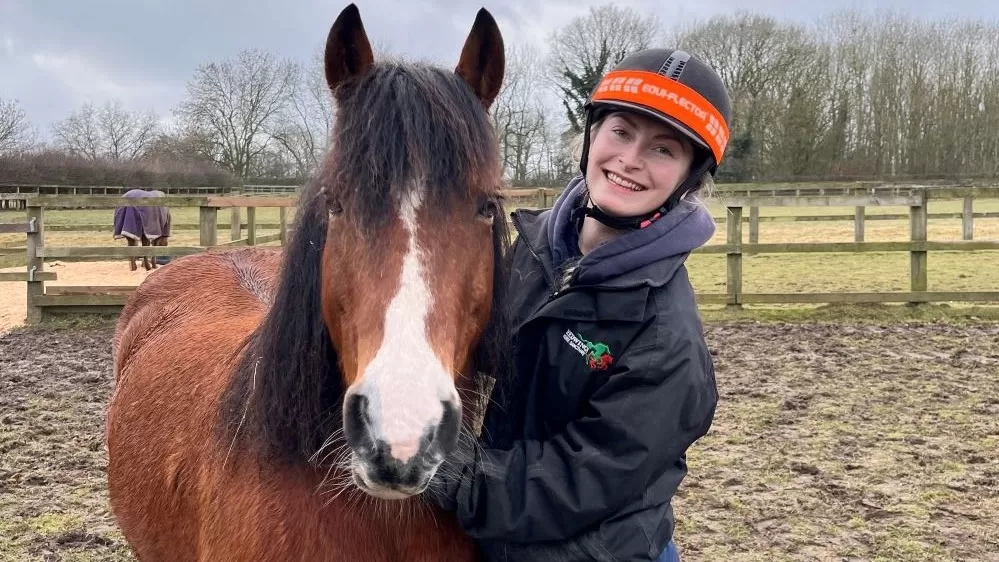 Chestnut pony Thaxton stands in his field, looking at the camera, while being cuddled by Equine Veterinary Nursing Apprentice Lizzy.