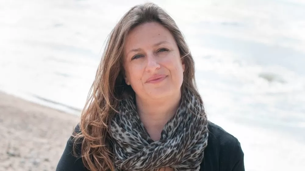 Author Elly Griffiths smiles at the camera while standing on a beach with the sea in the background.