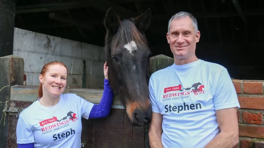 Two supporters wearing Redwings Horse Sanctuary running tops stand next to a stable with a horse poking their head over the top.