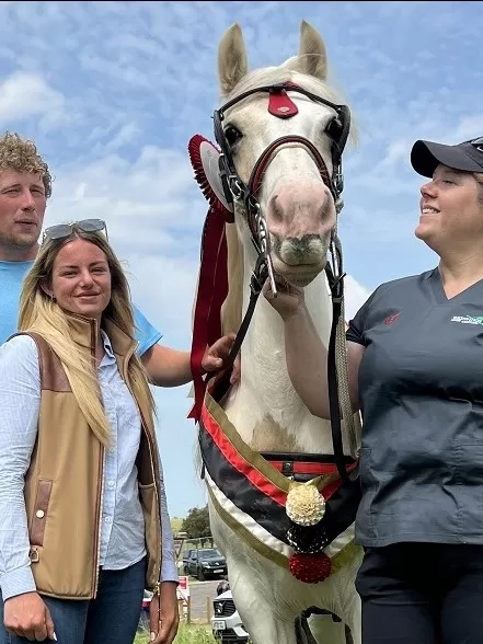 A brown and white pony wearing a winner's sash stands next to Redwings vet Nicola and their owners.